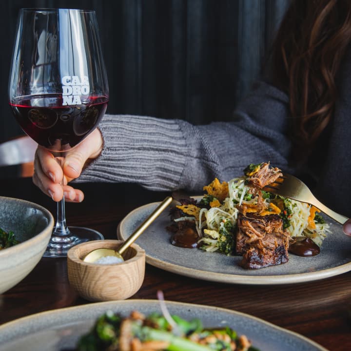 A hand holds a Cardrona-branded wine glass filled with red wine beside a plated meat dish with slaw and greens.