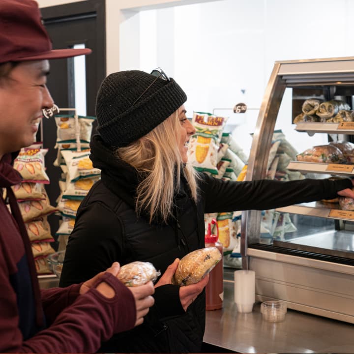 Two people in ski gear smile while selecting wrapped rolls and pastries from a self-serve display case.