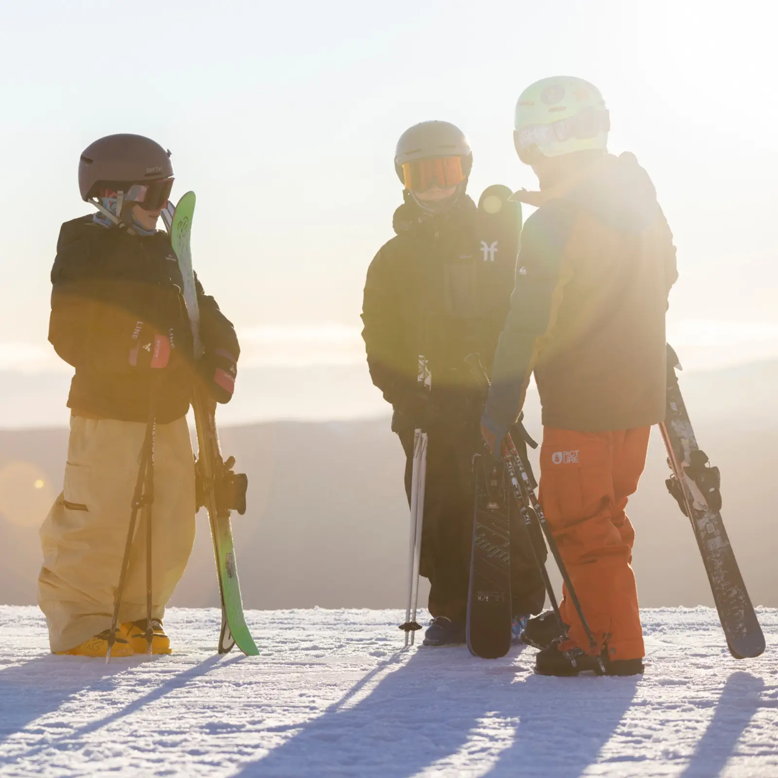 Three young skiers stand together on a snowy slope, holding their skis, bathed in warm golden sunlight with a misty mountain landscape behind them.