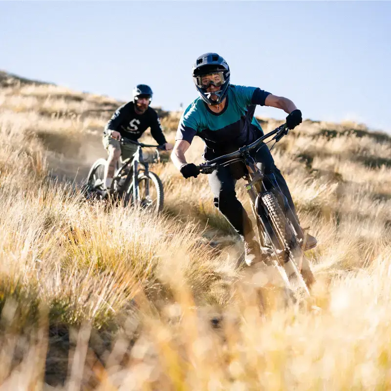 Two mountain bikers ride down a track on a blue sky summers day wearing t-shirts and full head helmets.