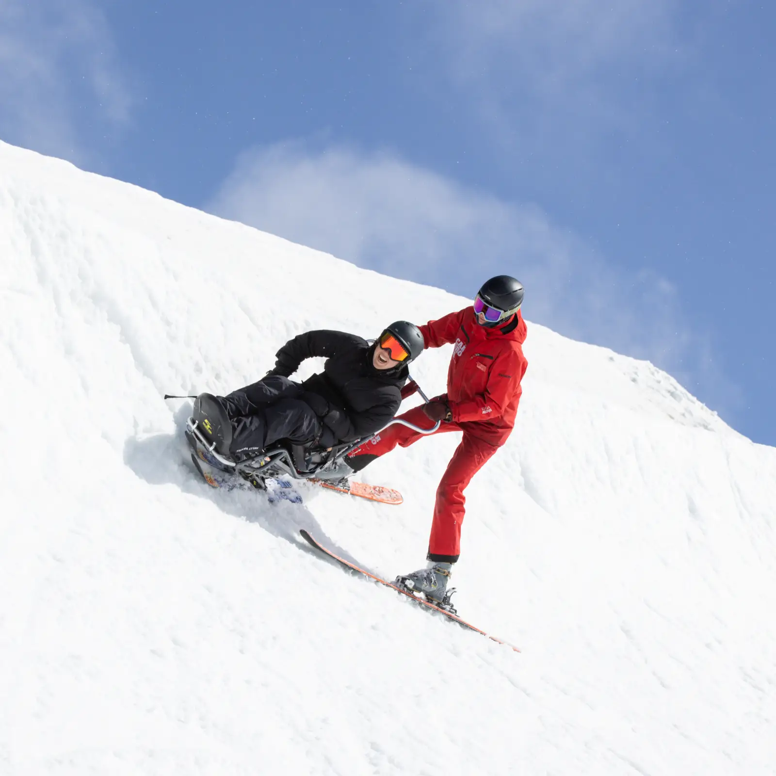 A sit-ski rider is guided downhill by an instructor in red on a snowy slope.