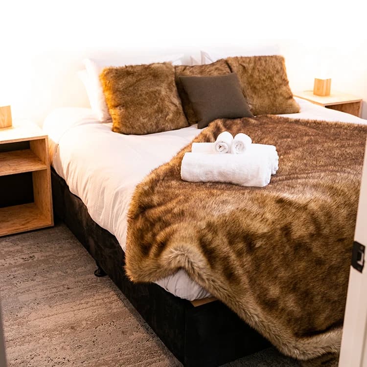A cosy bed with white sheets and brown fur throw and cushions.