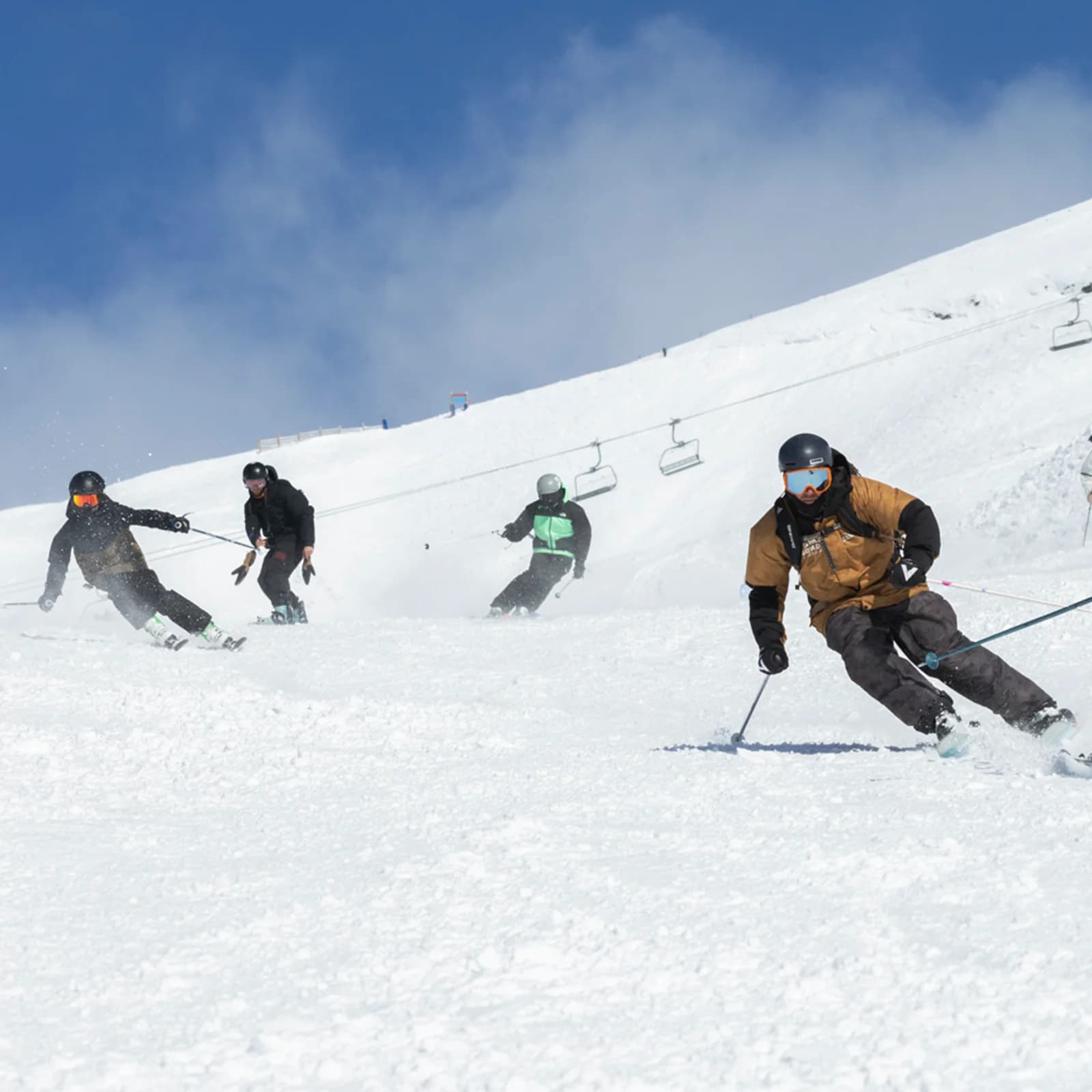 Four skiers descend a snowy mountain slope under a clear blue sky, with a ski lift in the background.