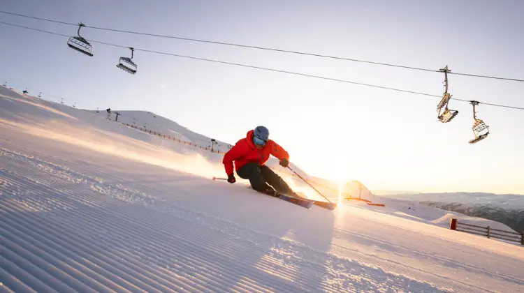 A skier in a red jacket carves down a freshly groomed slope at sunrise, with chairlifts overhead and snowy mountains in the background.