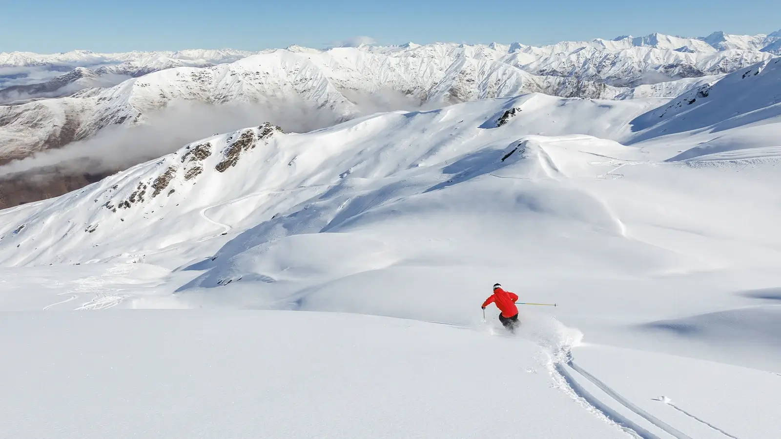 A skier in a red jacket carves through fresh powder on a vast snowy slope with rugged mountains stretching across the horizon.