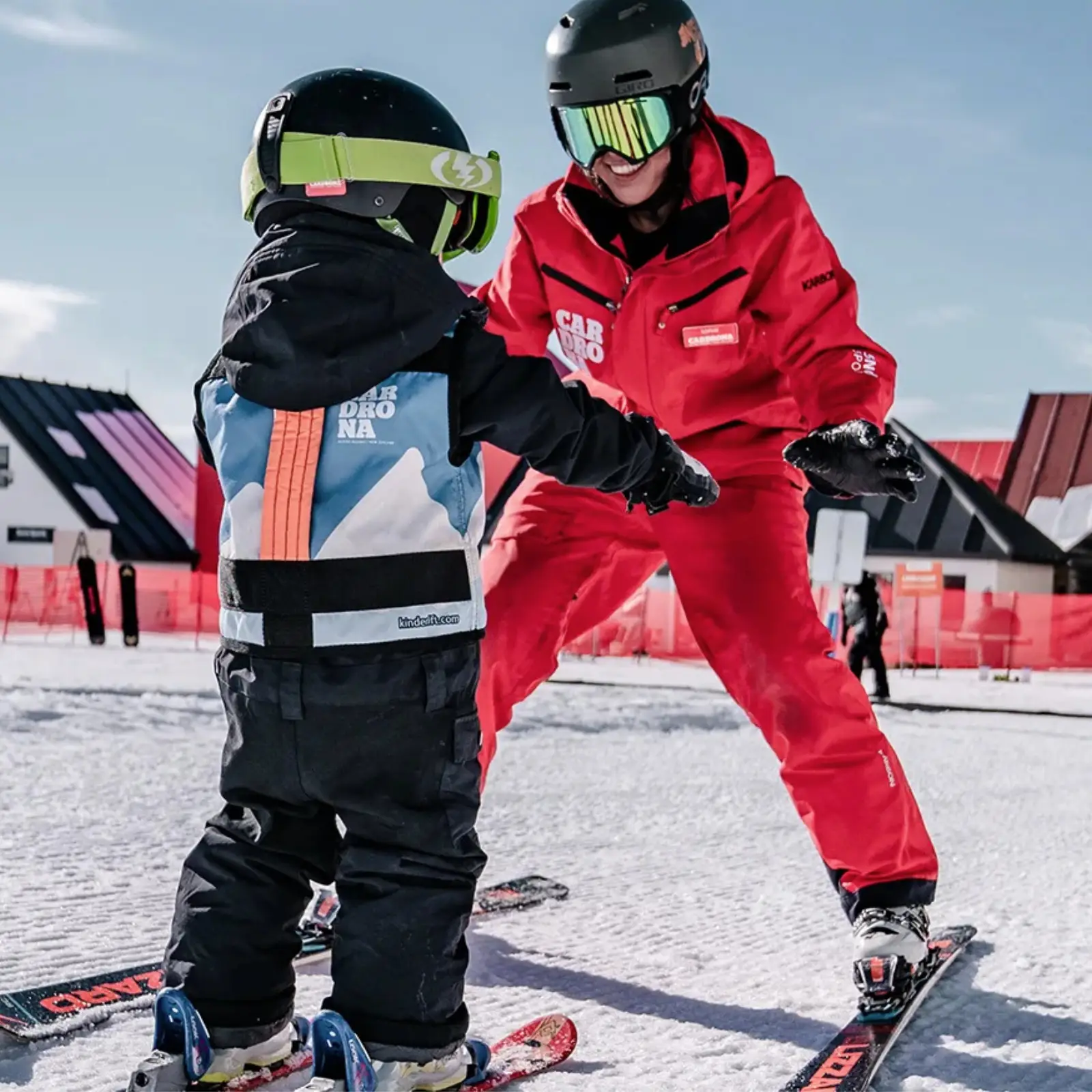A ski instructor dressed in Cardrona branded red ski clothes helps a young toddler standing on skis.