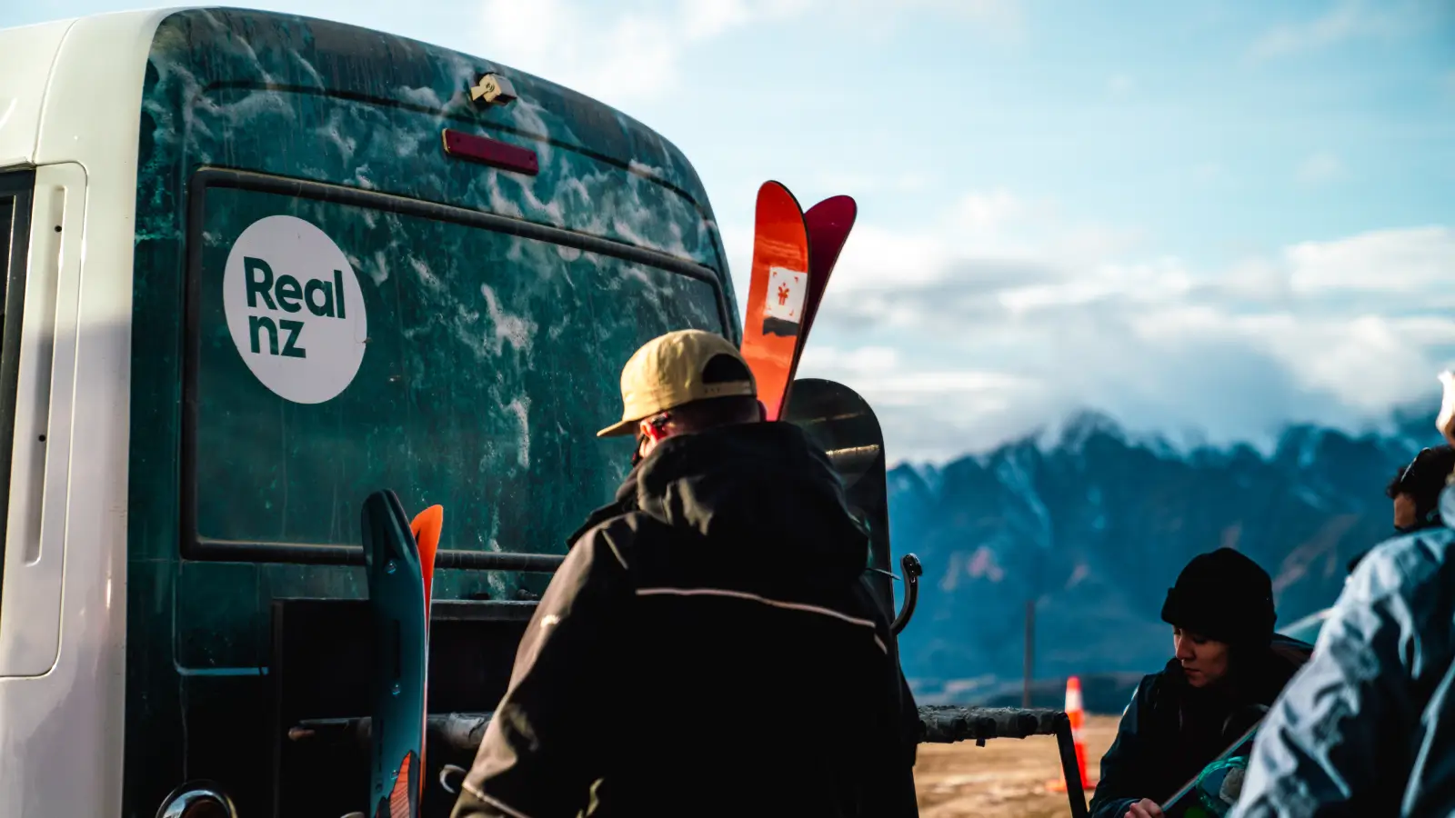 A man holding skis stands at the back of a RealNZ branded bus, ready to load the gear. Snowy mountains can be seen in the background.