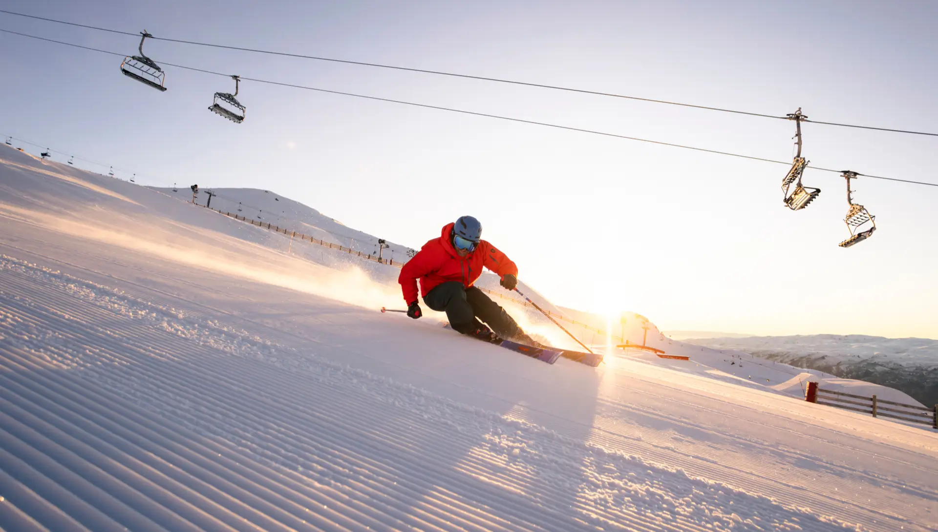 A skier in a red jacket carves down a freshly groomed slope at sunrise, with chairlifts overhead and snowy mountains in the background.
