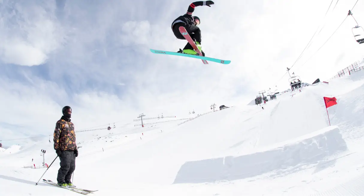 A skier launches high off a park jump with a teal board visible beneath them, while a coach watches from below.