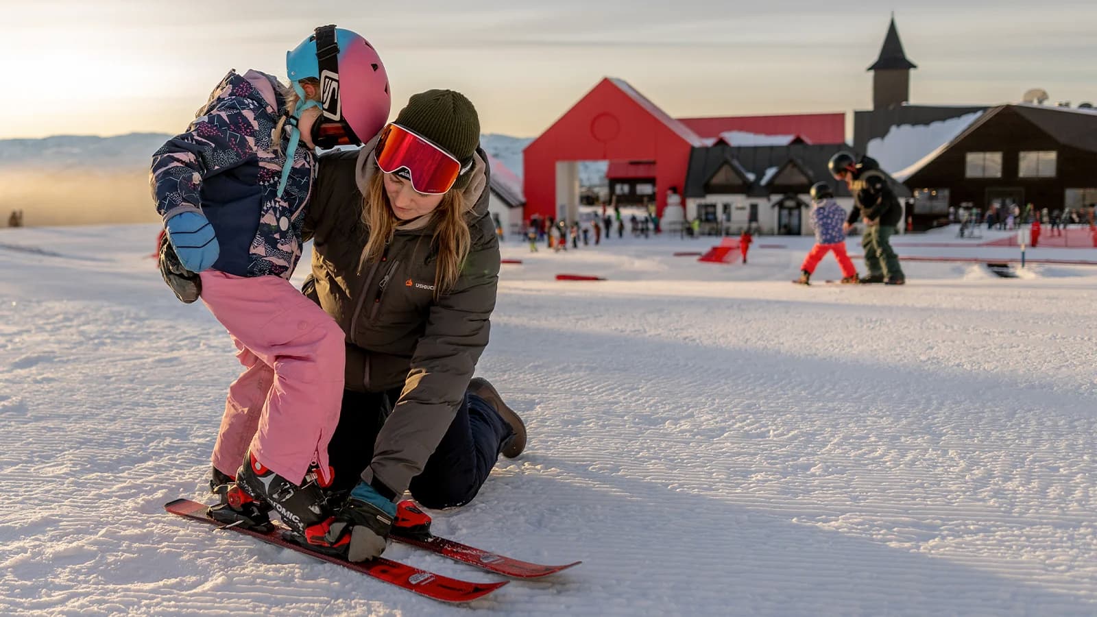 A parent helps their child strap into ski gear at the base of the Cardrona mountain. The sun is setting behind the lodge, casting a warm glow over the scene, while families enjoy the winter activities nearby.