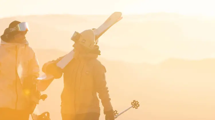 Two friends talk as they carry their ski and snowboard gear towards the bottom of the ski chairlift as the sunrises behind them.