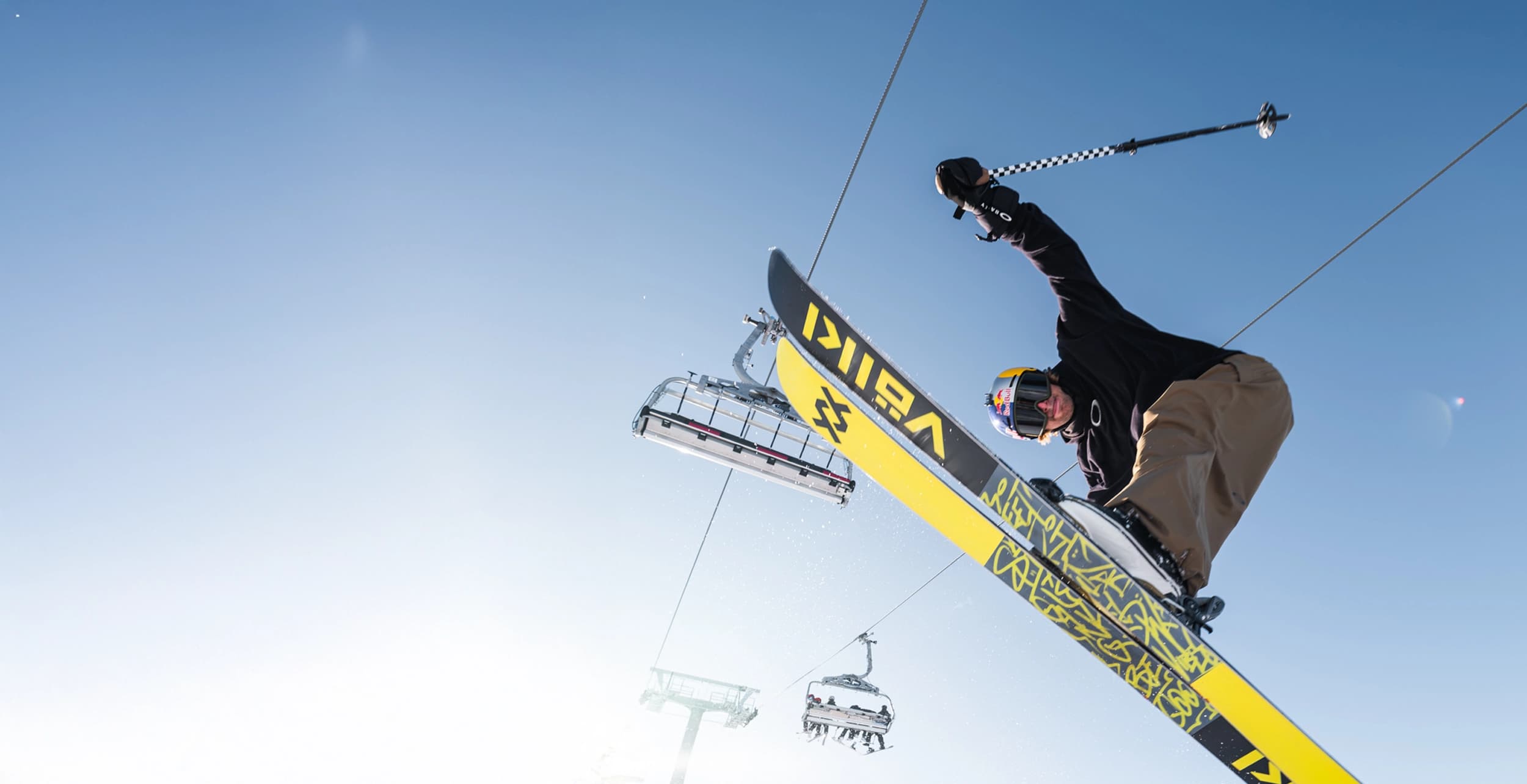 A skier performs an aerial trick under a clear blue sky, with a ski lift and mountain backdrop visible in the background.